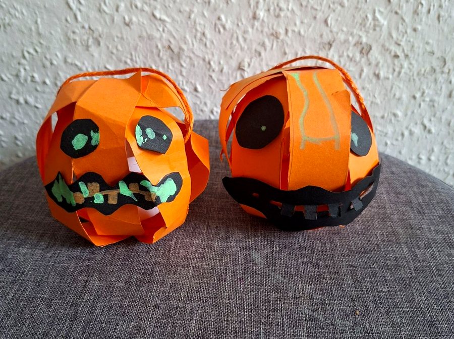 Zwei Kürbis-Teelichthalter aus orangefarbenen Papierstreifen mit aufgeklebten Augen und Mündern aus schwarzem Papier